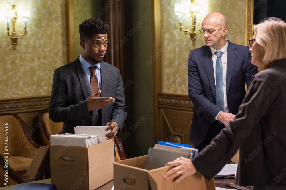 Poster young lawyer explaining something to one of mature colleagues while taking folder with papers out of