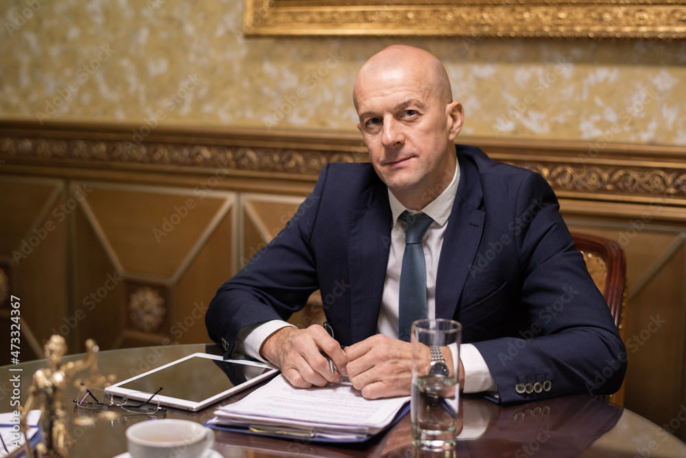 Poster mature white collar worker in elegant suit sitting by table in boarddroom and looking at camera whil