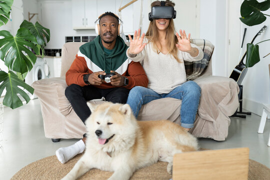 Smiling woman wearing virtual reality headset by man playing video game in front of dog at home