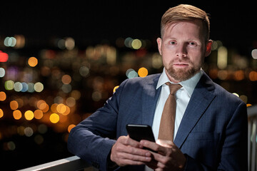 Young serious businessman with smartphone standing on balcony against night lights and looking at camera