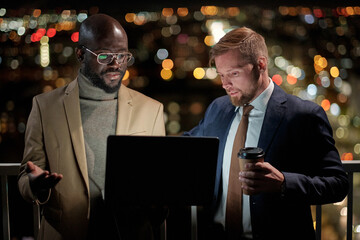 Contemporary businessmen looking at laptop screen during presentation or work planning at night meeting