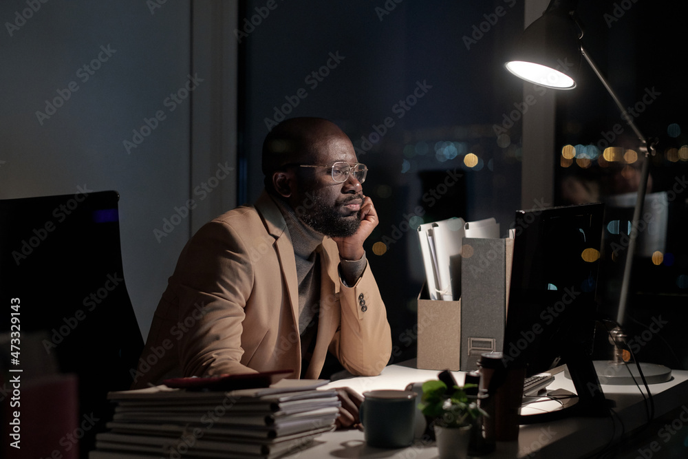 Poster African businessman sirring by desk in front of computer monitor in office while working late at night