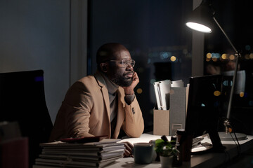 African businessman sirring by desk in front of computer monitor in office while working late at night