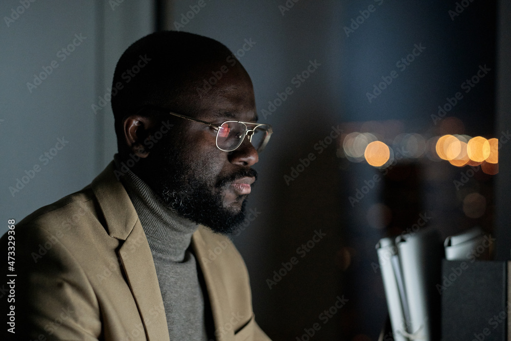 Poster young elegant businessman in formalwear and eyeglasses working in front of computer in office at nig