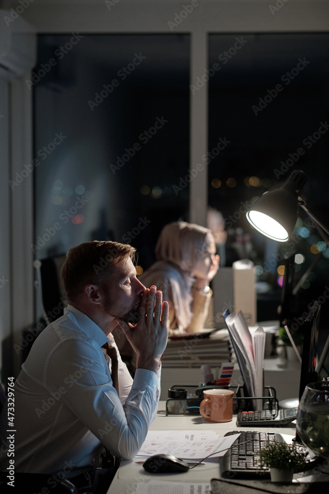 Poster tired businessman with his hands put together by face sitting in front of computer monitor against f
