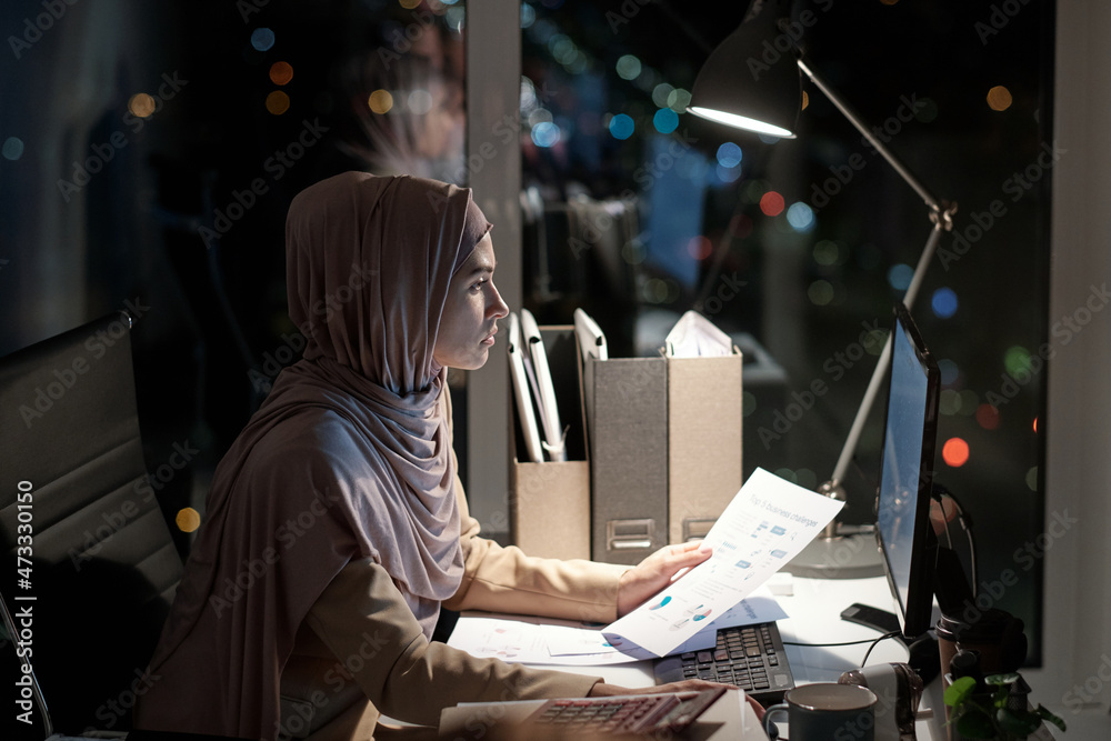 Poster young businesswoman with paper sitting in front of computer and looking through online data on scree