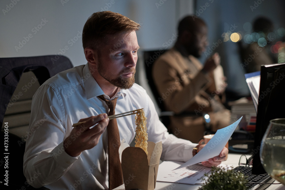 Poster busy white collar worker with paper eating chinese wok and looking at computer screen in office at n