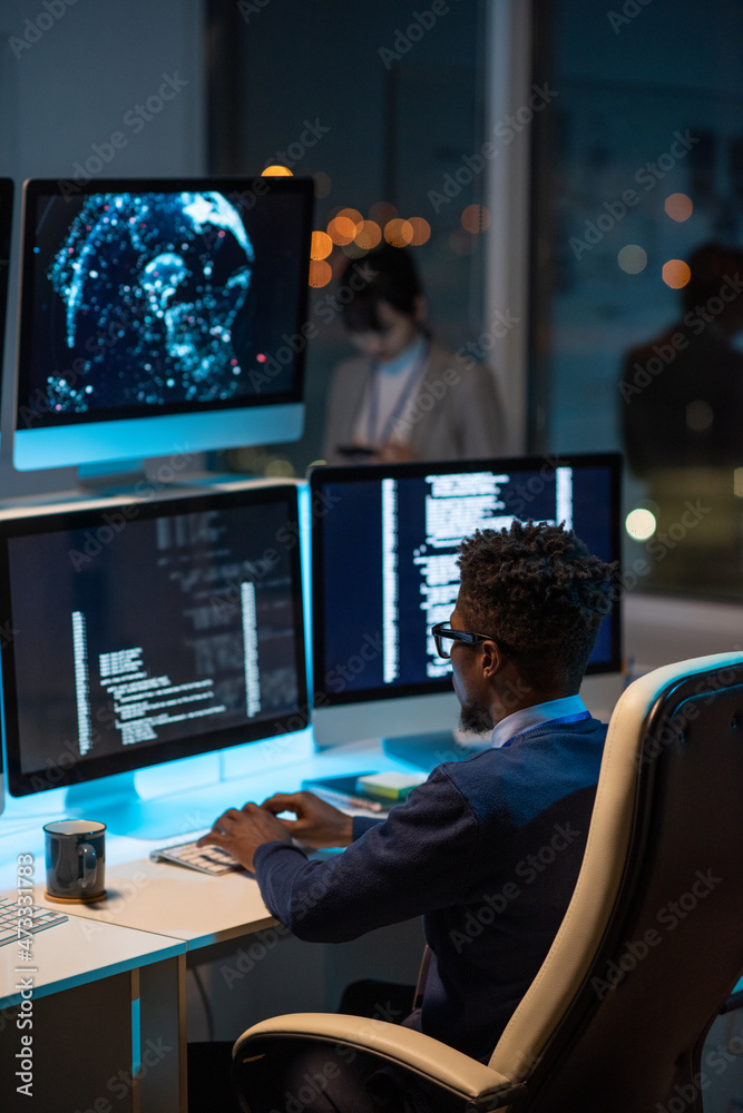 Aufkleber Young African developer sitting in armchair by desk and typing while looking at coded data on computer screen