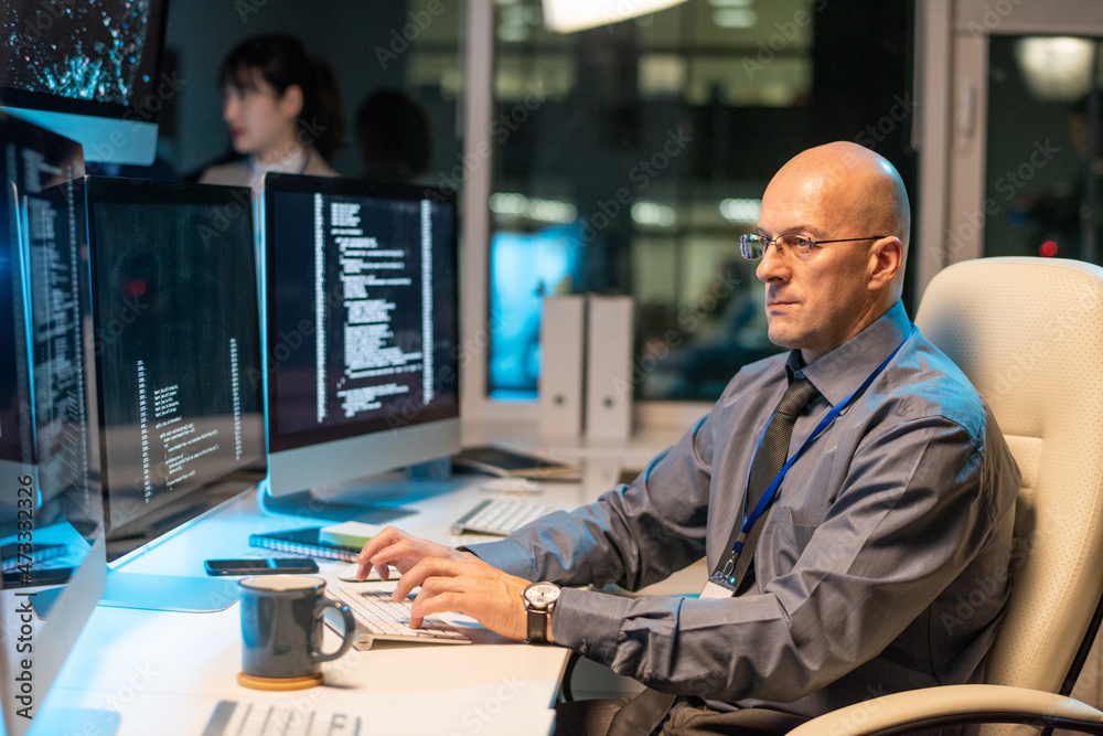 Poster Serious bald businessman looking through data on computer screen while sitting in front of monitor by desk