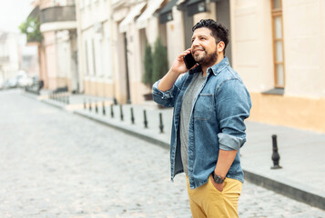 Young cheerful hispanic man in jeans jacket talking on phone on foggy european street