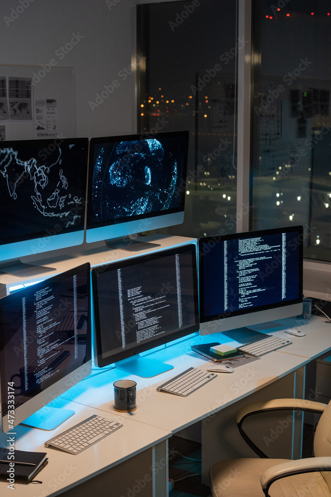 Fototapete Group of computer monitors with coded data standing in row on desks in empty office of programming company at night time