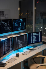 Group of computer monitors with coded data standing in row on desks in empty office of programming company at night time
