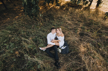 A young, smiling groom and a cute blonde bride with curly hair in a white dress are sitting on a rug on the yellow grass in the fall in nature and hugging. Wedding photo of the newlyweds, top view. - Powered by Adobe