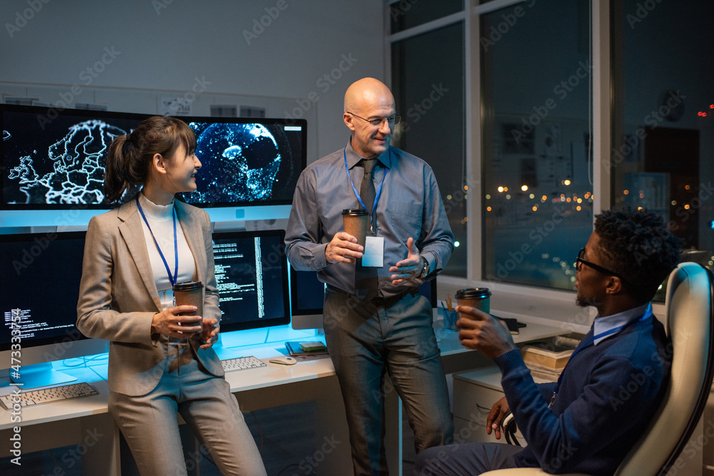 Poster Three happy intercultural colleagues with drinks chatting at break by workplace with computer monitors