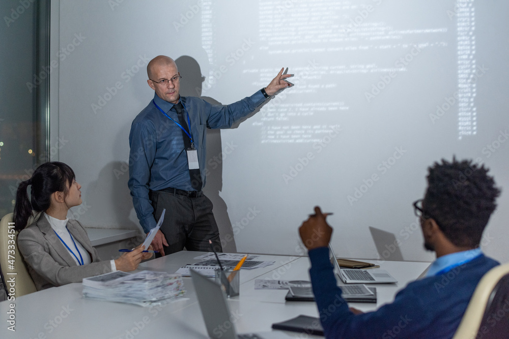 Poster Mature coach pointing at interactive whiteboard while explaining data to young intercultural developers at seminar