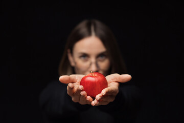 A smiling woman wearing glasses holding a red apple in palms show it to camera, with red lips, over a black background. Focus on fruit