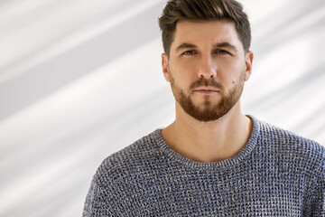 Obraz premium Photo portrait of a handsome man with a fashionable hairstyle and beard on a white background with shadows. A serious man with a stern look is wearing a gray sweater. Free space for text