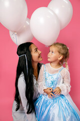 Young happy mother admiring her adorable daughter birthday girl with white balloons on a pink background. Cheerful brunette mom and her cute daughter are dressed in blue dresses