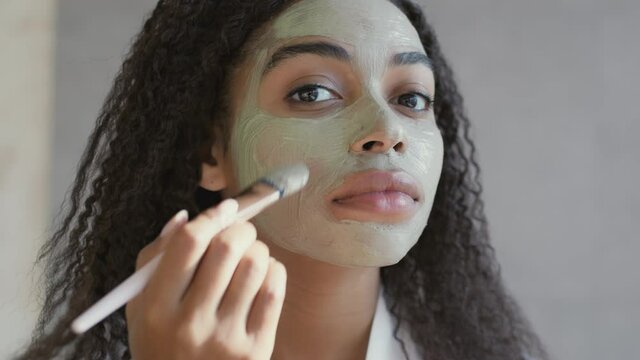 Home beauty procedures. Mirror pov of young african american woman applying pampering natural clay mask on her face