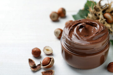 Glass jar with tasty chocolate spread and hazelnuts on white wooden table, space for text