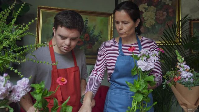 Young male florist with Down syndrome working with help of mentoring colleague indoors in flower shop.