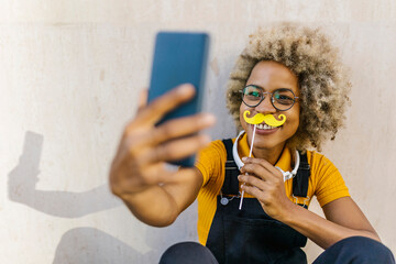 Woman holding fake mustache and taking selfie through smart phone in front of wall