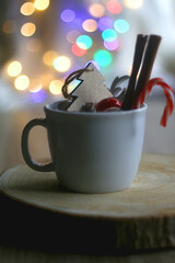 Mug filled with candy cane, cinnamon sticks and various Christmas decorations. Colorful bokeh lights in the background. Selective focus.