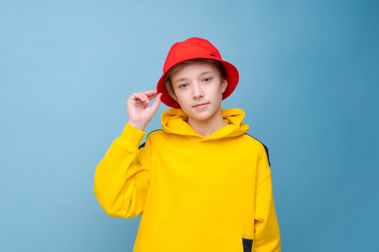 Rapper. City style. Model shooting. Happy hipster guy in yellow sweatshirt and red hat isolated blue background. Caucasian teenager posing in studio