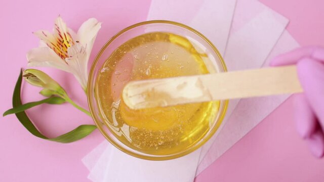 Liquid yellow sugar paste, wooden spatula and depilatory strips on a pink background. A master in pink gloves stir the sugar paste before removing unwanted hair. Sugaring.Depilation. Epilation.Beauty.