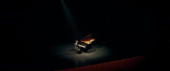 WIDE HIGH ANGLE Young 20s aspiring musician playing grand piano on a stage of a huge concert hall. Shot with 2x anamorphic lens