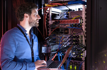 Male IT professional looking away while holding laptop by server rack in data center