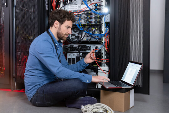 Male IT professional holding patch cord cable while working on laptop in server room