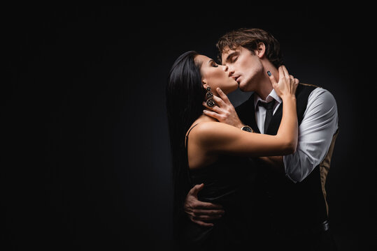man in vest and shirt kissing girlfriend in silk slip dress on black.