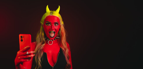 red demon girl with yellow eyes and yellow horns and hair wearing necklaces with spikes and black outfit with zipper on dark background holding a cellphone looking to the camera with her mouth open