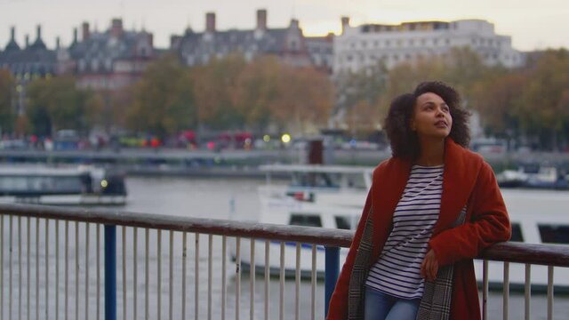 Woman wearing winter coat leaning on railings by river Thames in London at dusk - shot in slow motion
