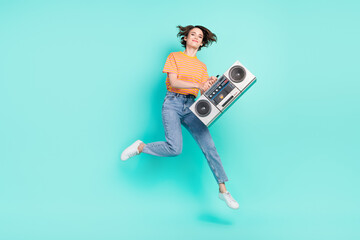Fototapeta na wymiar Photo of adorable funky young lady wear orange t-shirt jumping high holding boom box smiling isolated turquoise color background