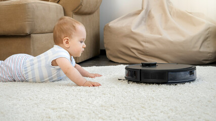 Little baby boy and robot vacuum cleaner approaching and getting closer on carpet in living room....