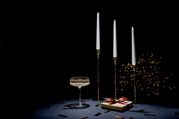 Three gold candlesticks with candles on a black background, next to a gift box and a glass of champagne