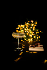 A glass of champagne on a black background with lights, a gift box lies nearby