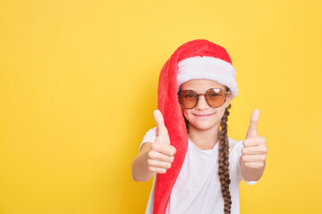 Fototapeta na wymiar cheerful teenage girl in sunglasses and red santa hat shows thumbs up on yellow background, copy space