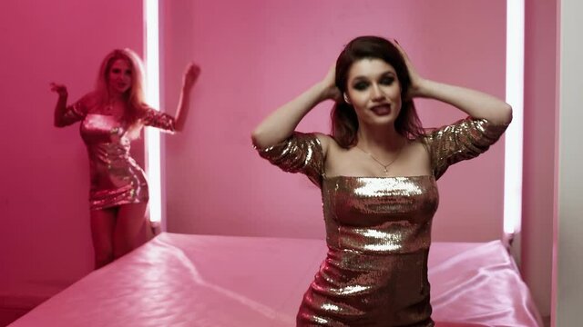 Two beautiful European girls in gold dresses dance and sing song for video clip in a studio with a bed and a pink background