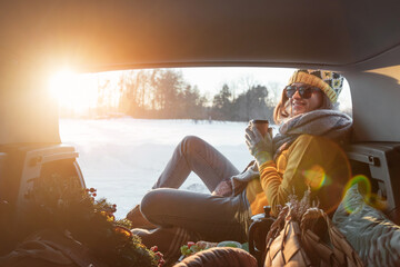 Woman with coffee in hands is sitting in car trunk and has winter picnic