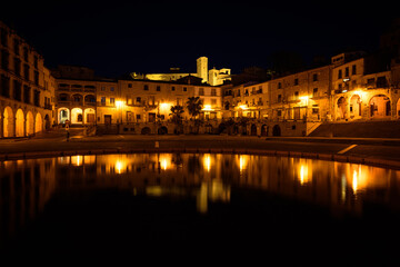 Fototapeta na wymiar City landscape of Trujillo old town with its old stone buildings reflected in the fountain and the fortress palace of Chaves and Santiago Gate in the background at night, Caceres, Extremadura, Spain