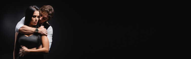 man in vest and white shirt hugging brunette girlfriend in silk slip dress on black, banner.