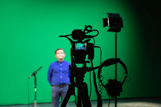 Videography of a boy in a blue shirt in the studio on a green background. Chromakey technology. A microphone on a stand, a video camera on a tripod and lighting equipment.