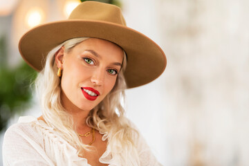 Fashionable blond haired woman wearing hat and red lipstick while looking at camera and smiling