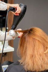 Drying hair in the hair studio. Female hairdresser stylist dries hair with a hairdryer and round brush red hair of a woman in a beauty salon.