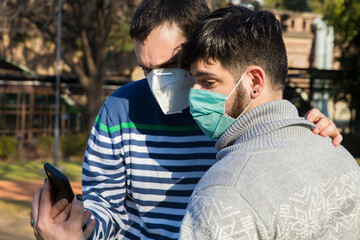 Young gay couple wearing protective mask carefully reading social media news on smartphone...
