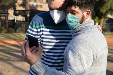 Young gay couple wearing protective mask carefully reading social media news on smartphone...