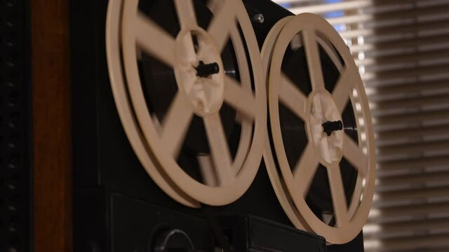 On a reel-to-reel tape recorder, reels are spinning against the background of a window with shutters, tape is spinning, close-up, selective focus. Sound recorder, high quality sound, spy tape recorder